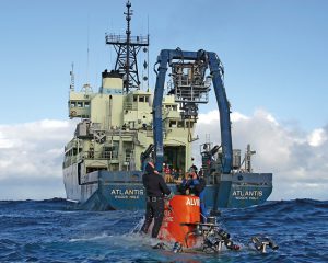Research Vessel Atlantis and the human-occupied vehicle Alvin are shown. Divers meet the sub when it surfaces to attach it to a hoist rope and bring it back on board (Image Woods Hole Oceanographic Institution). 