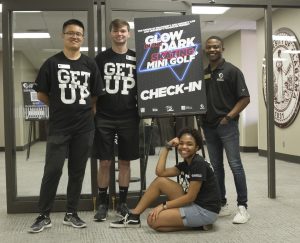 Four university students pose for a photo near an advertisement for a campus event.