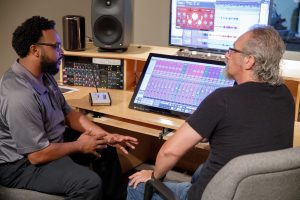 Two men sit at a table in a recording studio. 