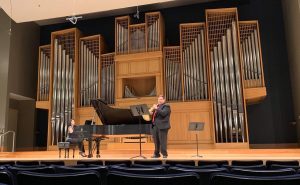 Matthew Meadows plays the French horn on a stage with a pianist.