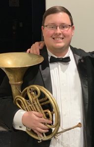 Matthew Meadows holds a French horn and wears a tuxedo.