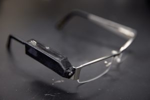 A pair of eyeglasses, with a prototype of the ingestion monitor attached to one leg, sit on a campus lab bench. 
