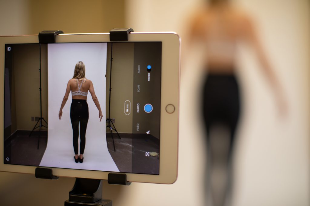 A woman in work-out clothes stands still while a tablet computer captures an image of her, shown on screen.
