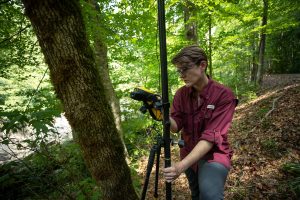 A researcher stands using s GPS device on a tripod in the forest.