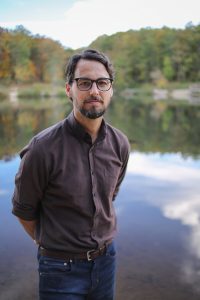 A man in jeans and button-down shirt poses with hands behind his back in front of a lake.