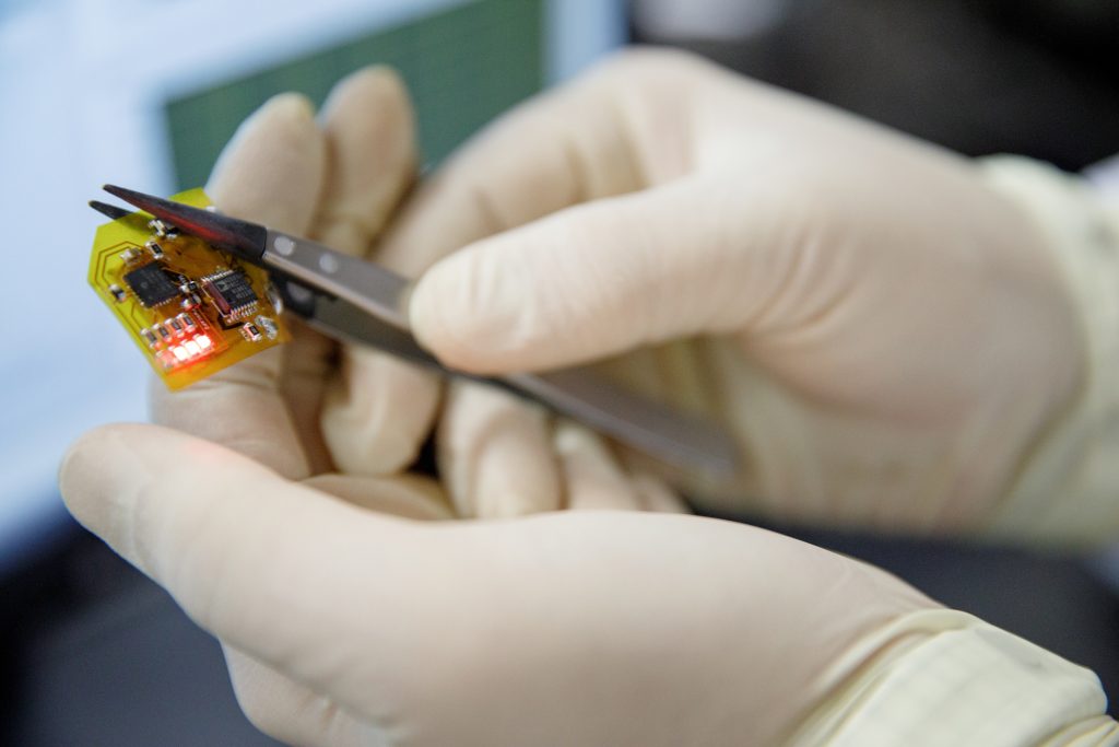 A pair of gloved hands holds a small electronic chip in tweezers