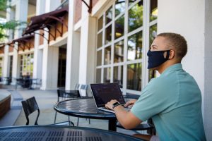 A man wearing a mask uses a laptop at outdoor table