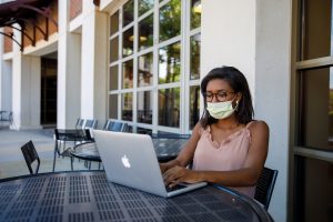 A woman wearing a face mask uses a lap top at a patio table at The University of Alabama