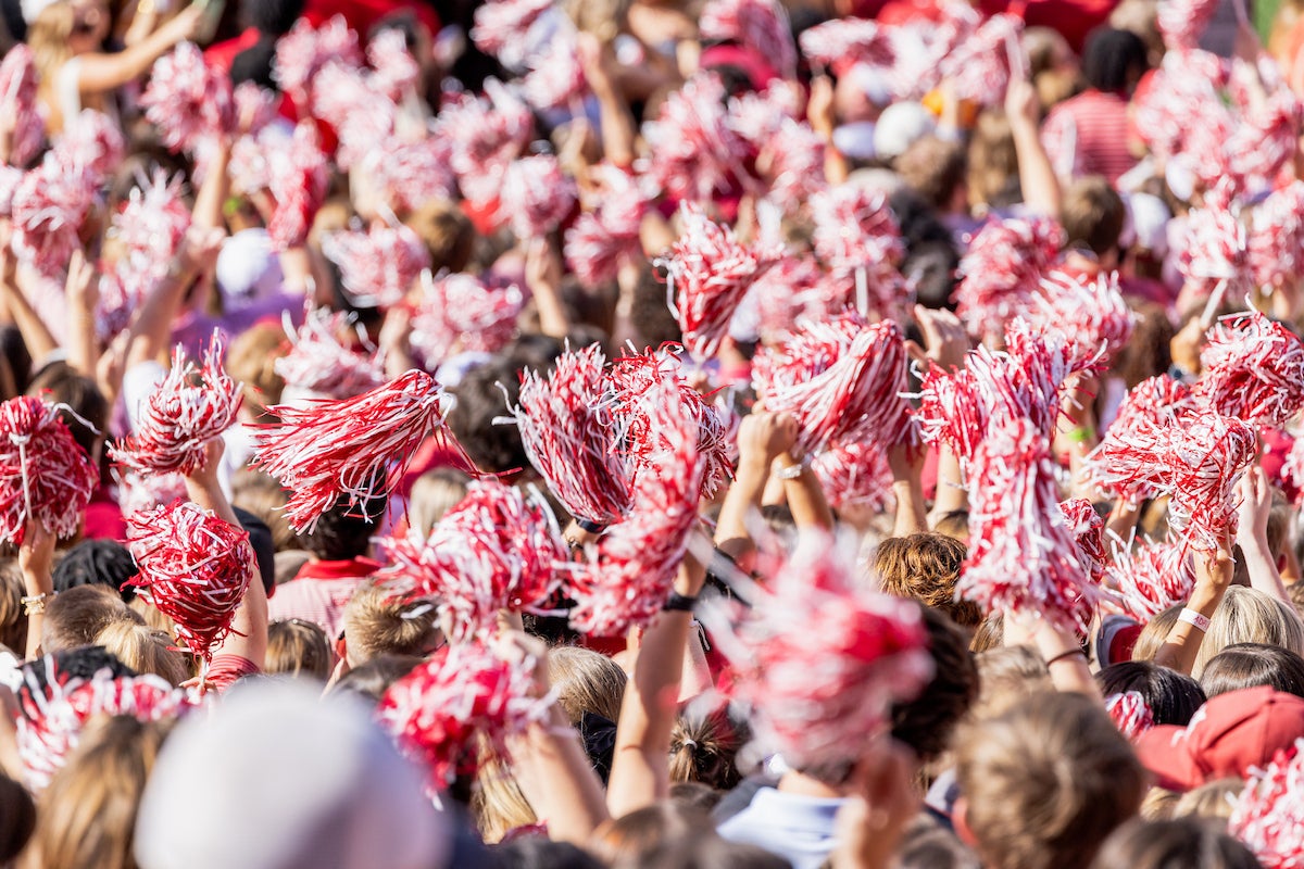 A packed stadium crowd waves red and white shakers in the air during a University of Alabama football game.