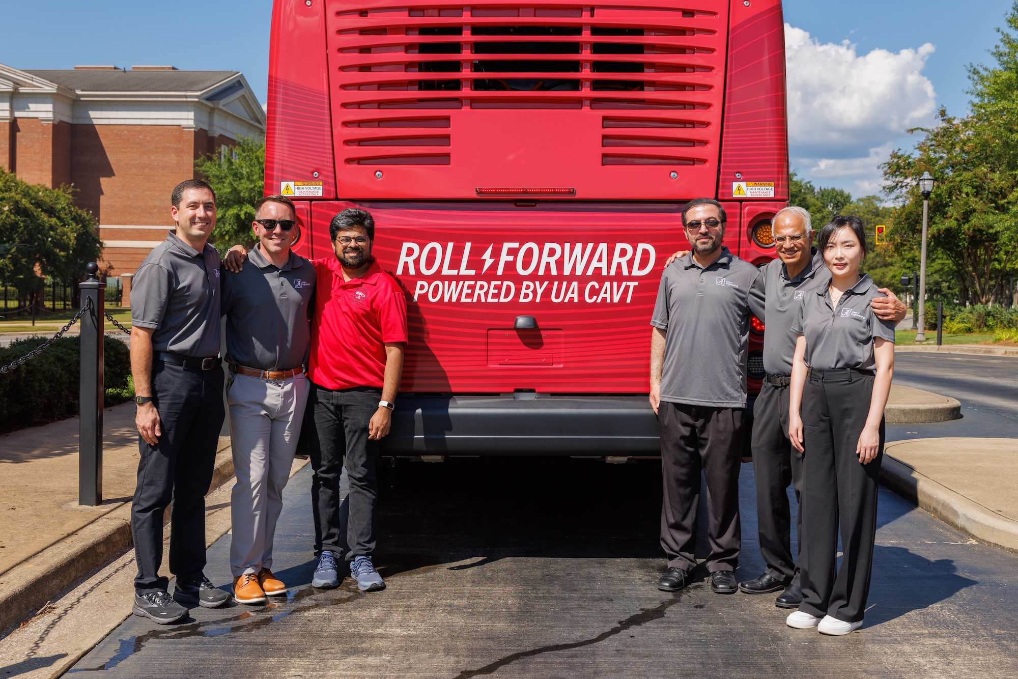 Six members of the UA research team stand at the back of a red bus lettered 