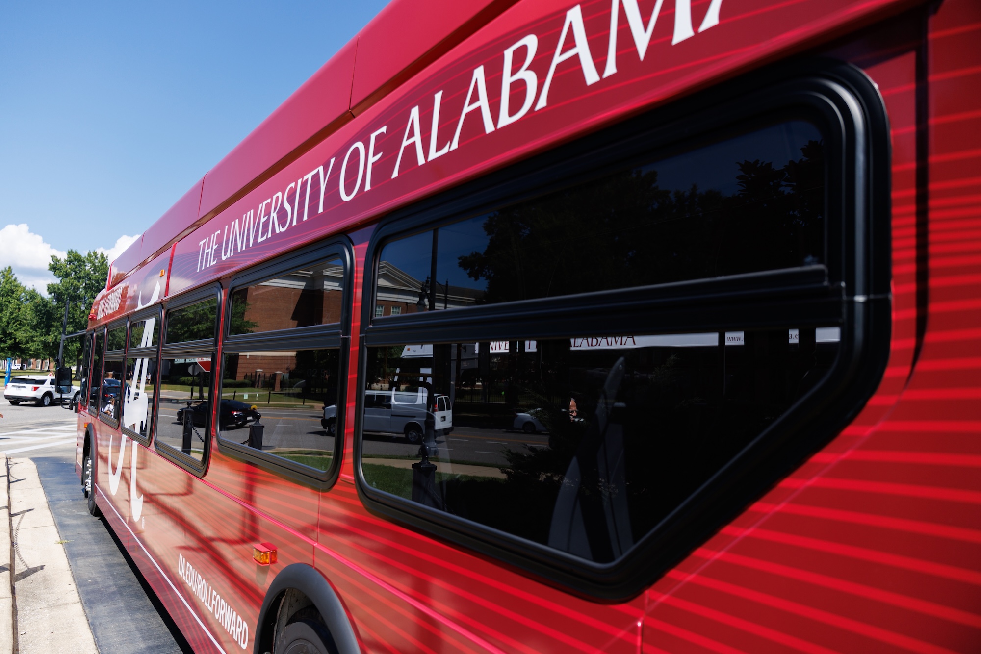 The side of a red University of Alabama bus, campus buildings reflected in its windows.